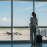 Młoda kobieta czeka z walizką przy oknie lotniska, za oknem samolot przygotowujący się do boardingowania