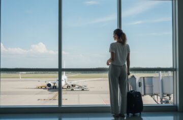 Młoda kobieta czeka z walizką przy oknie lotniska, za oknem samolot przygotowujący się do boardingowania