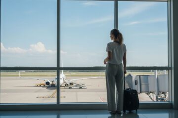Młoda kobieta czeka z walizką przy oknie lotniska, za oknem samolot przygotowujący się do boardingowania