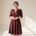 Pewna siebie niska kobieta plus size w sukience z dekoltem w serek pozująca na beżowym tle.