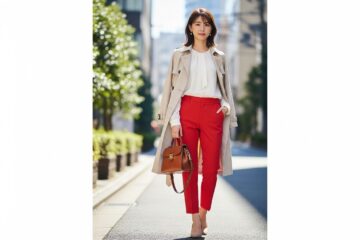Młoda kobieta na ulicy w czerwonych spodniach, białej bluzce i beżowym płaszczu, trzymająca małą torebkę.