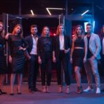 Grupa młodych osób w modnych strojach stoi przed klubem nocnym przy kolorowych neonach.