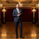 Mężczyzna w garniturze stoi w eleganckim lobby teatru, poprawiając spinki mankietowe.