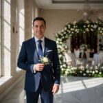 Mężczyzna w granatowym garniturze z kwiatem, stojący w eleganckim korytarzu ślubnym w naturalnym świetle